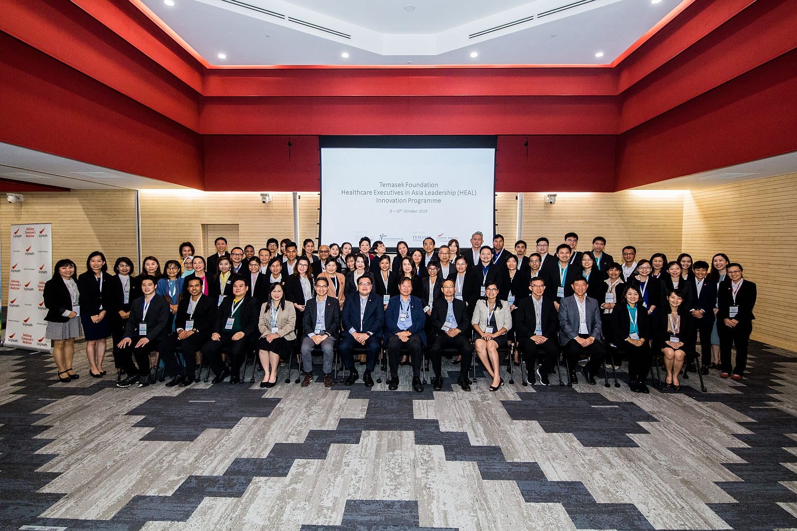 A large group of people in formal attire pose for a photo in front of a screen and red ceiling in a conference room.